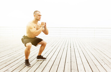 Muscular male athlete with naked torso doing squats exercise on the beach