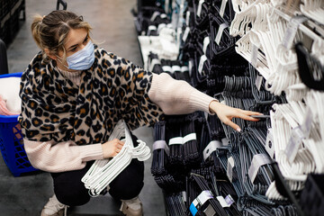 Young woman in medical mask crouches down and selects set of plastic hangers for organizing dressing room space in shopping center. New normal concept. Shopping in epidemic. Order in house.