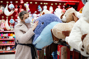 Pretty woman in mask and coat is standing next to huge box of stuffed toys and stroking large blue whale . Children's toy store. Shopping during the pandemic. Protective equipment.