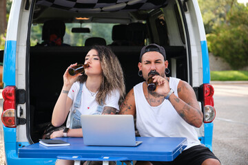 Young tattooed couple smile, drinking beer and using the laptop.