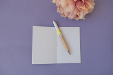 Cahier ouvert sur une page blanche sur un bureau mauve avec une fleur rose et un stylo pour un style féminin - obrazy, fototapety, plakaty