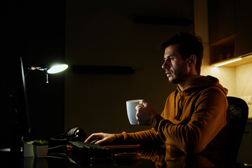 Obraz na płótnie Canvas Man works at home office workplace with cup of coffeee in his hand, use computer. Freelancer remotely working late. Emotional stress and burnout