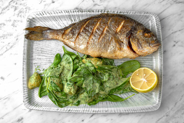Dorado grilled fish with spinach salad and cheese in a festive plate on a marble background. Restaurant banquet menu.