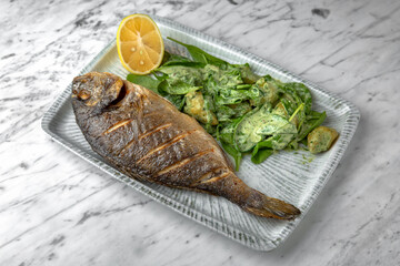 Dorado grilled fish with spinach salad and cheese in a festive plate on a marble background. Restaurant banquet menu.