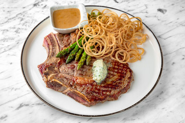 Big delicious juicy bone steak with onion rings in a festive plate on a marble background. Restaurant banquet menu.