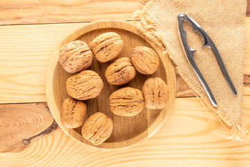Several ripe delicious walnuts with a round wooden tray and a metal nutcracker on a wooden table, close-up, top view.