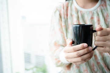Young Asian woman holding a coffee cup near the window, woman drinking a coffee. Happy woman drinking hot coffee in a mug in the morning close up with copyspace.
