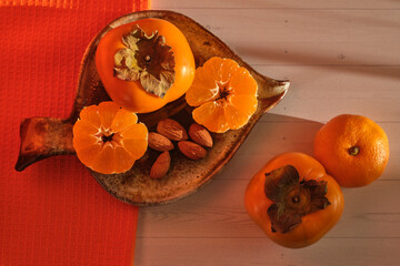 still life of tropical fruits: persimmons and tangerine cut in half and almonds on a clay saucer. Orange napkin on a 
light table background, healthy food concept