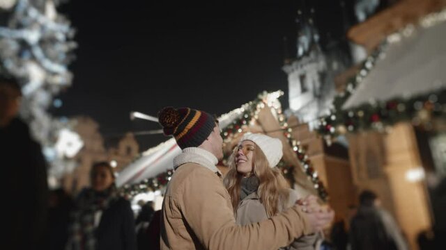 Touching moment during holiday season celebration. Beautiful scene of friends couple dancing slowly in a middle of christmas fare in european town.