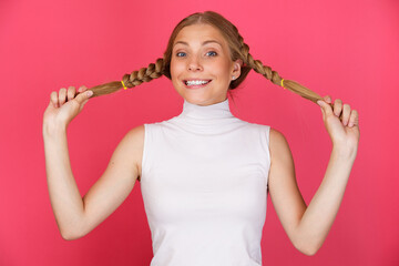 Portrait of cheerful smiling and winking woman holding long hair pigtails on pink background.