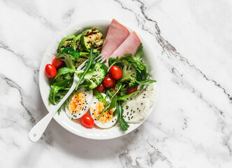 Delicious balanced breakfast, snack bowl - ham, boiled egg, grilled zucchini, green salad in one bowl on a light background, top view