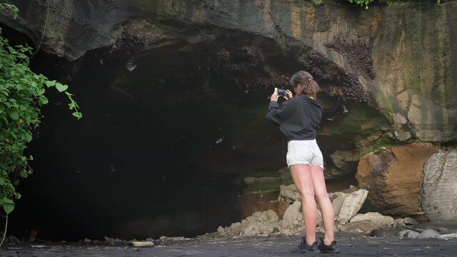 Girl stands in front of entrance to a large cave from which bats fly out and takes video on her phone. A woman in denim shorts records a video of bats on island of Bali in Indonesia. slow motion.