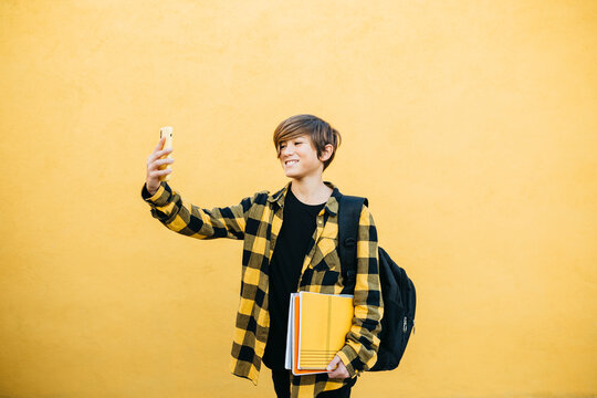 Portrait of preteenager smiling student boy with notebooks and backpack taking a selfie with smartphone in front of a yellow background