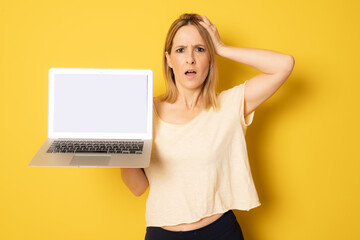 Confused woman puts her hands on the head showing laptop computer screen isolated on yellow background.