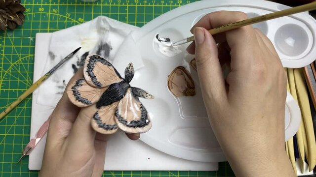 Artist holds brooch in form of butterfly with one hand, dips brush into palette with acrylic with other hand and applies paint to jewelry. Moth figurine made of clay. Decoration designer's workshop.