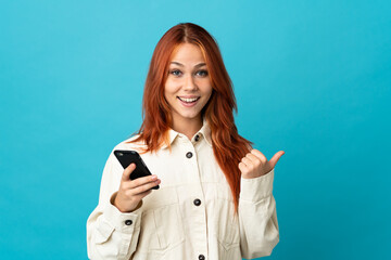 Teenager Russian girl isolated on blue background using mobile phone and pointing to the lateral