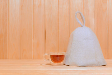 Glass mug with tea and hat is on wooden shelf in the bathhouse.