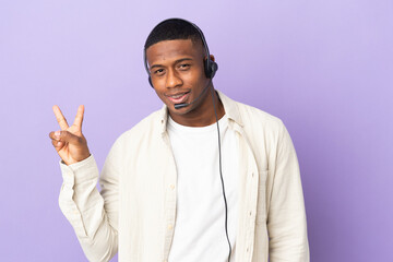 Telemarketer latin man working with a headset isolated on purple background smiling and showing victory sign