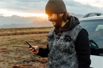 Caucasian man, traveler and adventurer with a beard and tattoos consulting his mobile phone leaning on his 4x4 car at sunset.