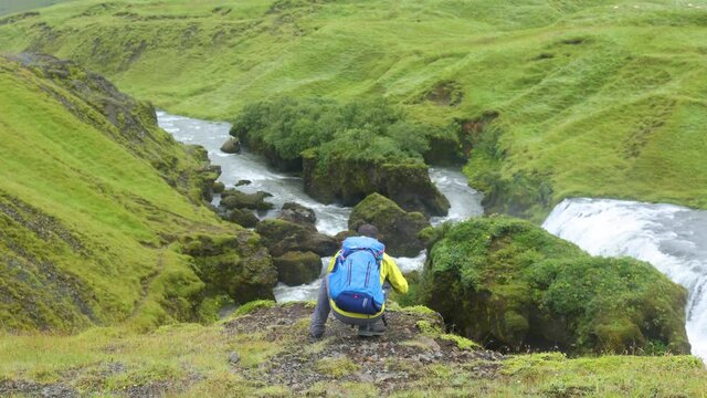 photographer is sitting on an edge of a cliff take pictures of the waterfall and streams in Iceland.  The picture taker is backpacking across the landscape enjoying the environment 