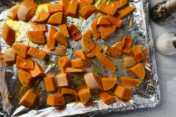 Raw butternut squash cut into pieces, on an oven tray covered with aluminum foil. Seasoned with...