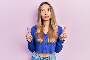 Beautiful hispanic woman wearing casual blue shirt pointing up looking sad and upset, indicating...