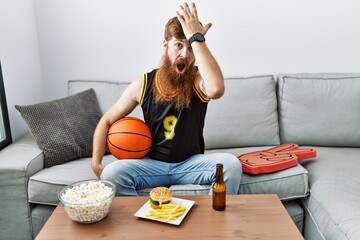 Caucasian man with long beard holding basketball ball cheering tv game surprised with hand on head...