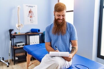 Fototapeta premium Young redhead man wearing physiotherapist uniform writing medical report at physiotherapy clinic