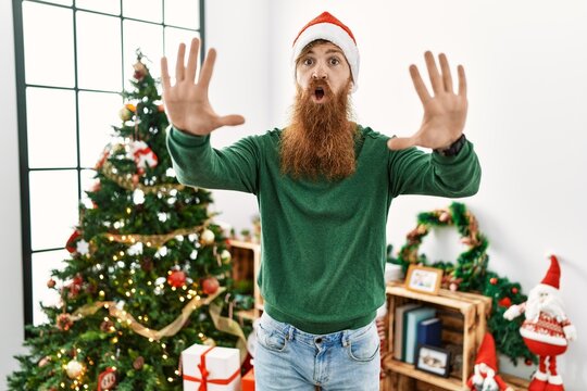 Redhead man with long beard wearing christmas hat by christmas tree doing stop gesture with hands palms, angry and frustration expression