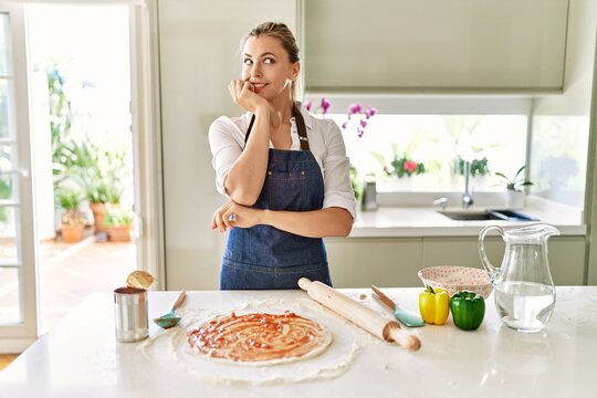 Beautiful blonde woman wearing apron cooking pizza looking stressed and nervous with hands on mouth biting nails. anxiety problem.