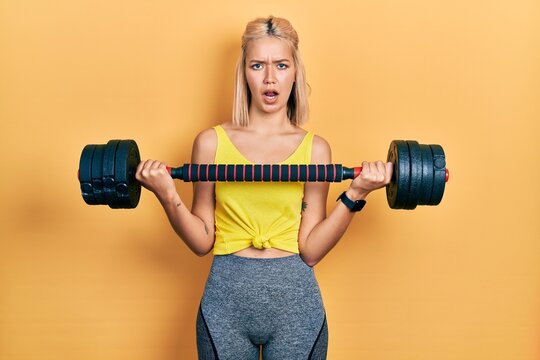 Beautiful blonde woman wearing sportswear using dumbbells in shock face, looking skeptical and sarcastic, surprised with open mouth