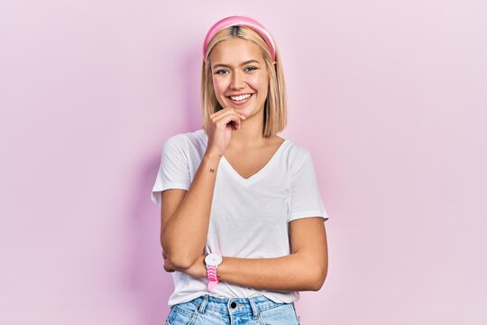 Beautiful blonde woman wearing casual white t shirt looking confident at the camera with smile with crossed arms and hand raised on chin. thinking positive.