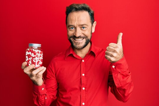 Middle age man holding pills smiling happy and positive, thumb up doing excellent and approval sign