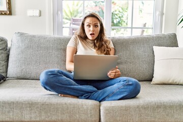 Beautiful young brunette woman sitting on the sofa using computer laptop at home afraid and shocked...