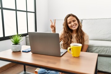 Beautiful hispanic woman using computer laptop at home smiling with happy face winking at the camera doing victory sign. number two.