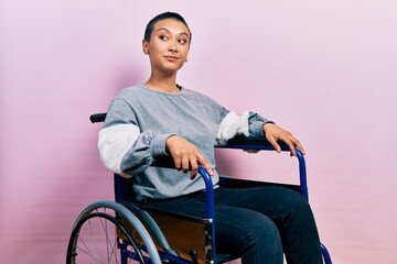 Beautiful hispanic woman with short hair sitting on wheelchair smiling looking to the side and...