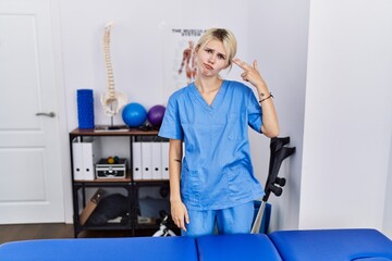 Young physiotherapist woman working at pain recovery clinic shooting and killing oneself pointing hand and fingers to head like gun, suicide gesture.