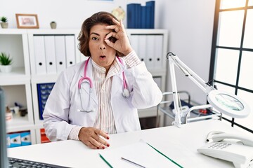 Middle age hispanic woman wearing doctor uniform and stethoscope at the clinic doing ok gesture shocked with surprised face, eye looking through fingers. unbelieving expression.