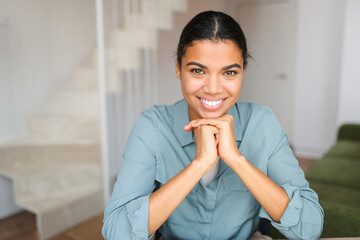 Fototapeta na wymiar Portrait of charming elegant woman with hands under chin smiling at the camera, sitting in the modern house, wearing jeans shirt