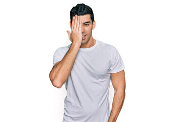Naklejka premium Handsome hispanic man wearing casual white t shirt covering one eye with hand, confident smile on face and surprise emotion.