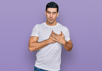 Handsome hispanic man wearing casual white t shirt smiling with hands on chest with closed eyes and grateful gesture on face. health concept.