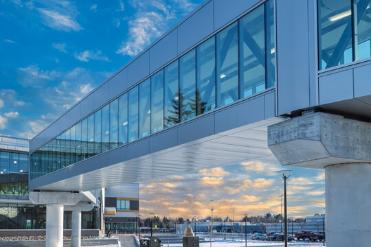 Exterior of metal and glass clad walkway supported on concrete piers between two buildings during daytime, blue and gold sky, nobody