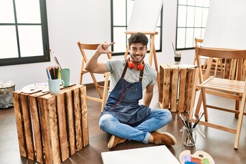 Young hispanic man sitting at art studio smiling and confident gesturing with hand doing small size sign with fingers looking and the camera. measure concept.