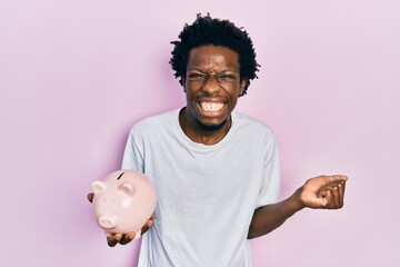 Young african american man holding piggy bank screaming proud, celebrating victory and success very...