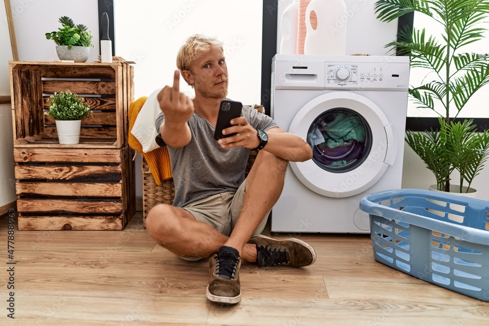 Poster young blond man doing laundry using smartphone showing middle finger, impolite and rude fuck off exp