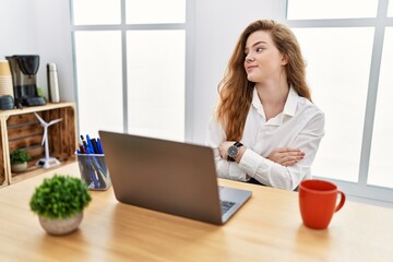 Young caucasian woman working at the office using computer laptop looking to the side with arms...