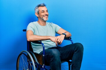 Handsome middle age man with grey hair sitting on wheelchair looking away to side with smile on...