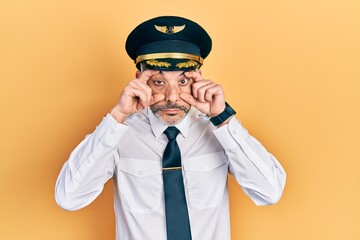 Handsome middle age man with grey hair wearing airplane pilot uniform trying to open eyes with...