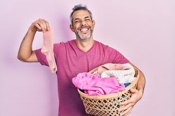 Handsome middle age man with grey hair holding laundry basket and dirty sock smiling and laughing...