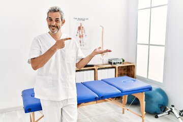 Middle age hispanic therapist man working at pain recovery clinic amazed and smiling to the camera while presenting with hand and pointing with finger.